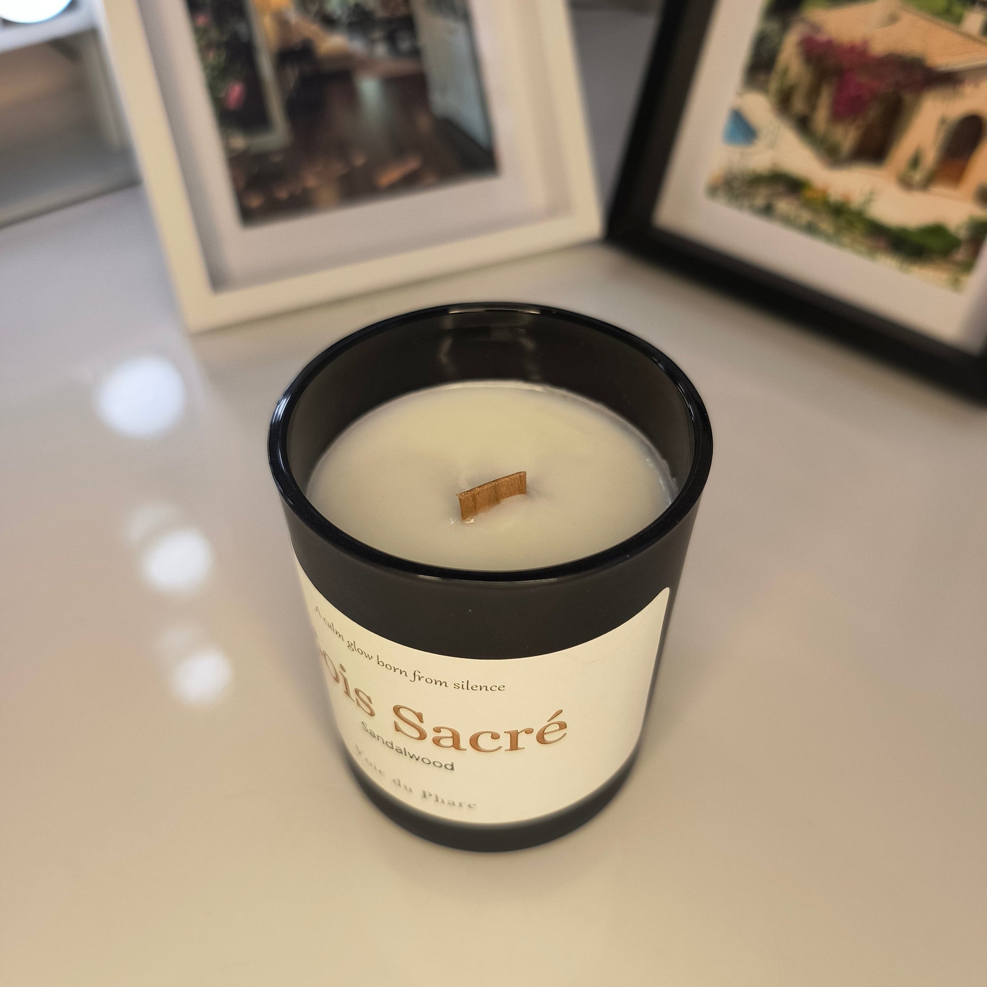 Candle in a black container on a surface with framed pictures in the background