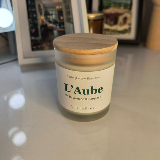 Jar of candle labeled 'L'Aube' on a reflective surface with a blurred background