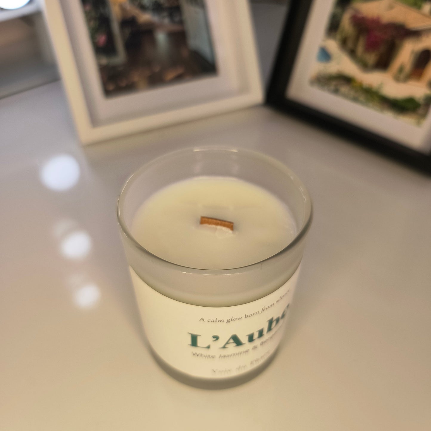 Candle with a label on a white surface, with framed pictures in the background
