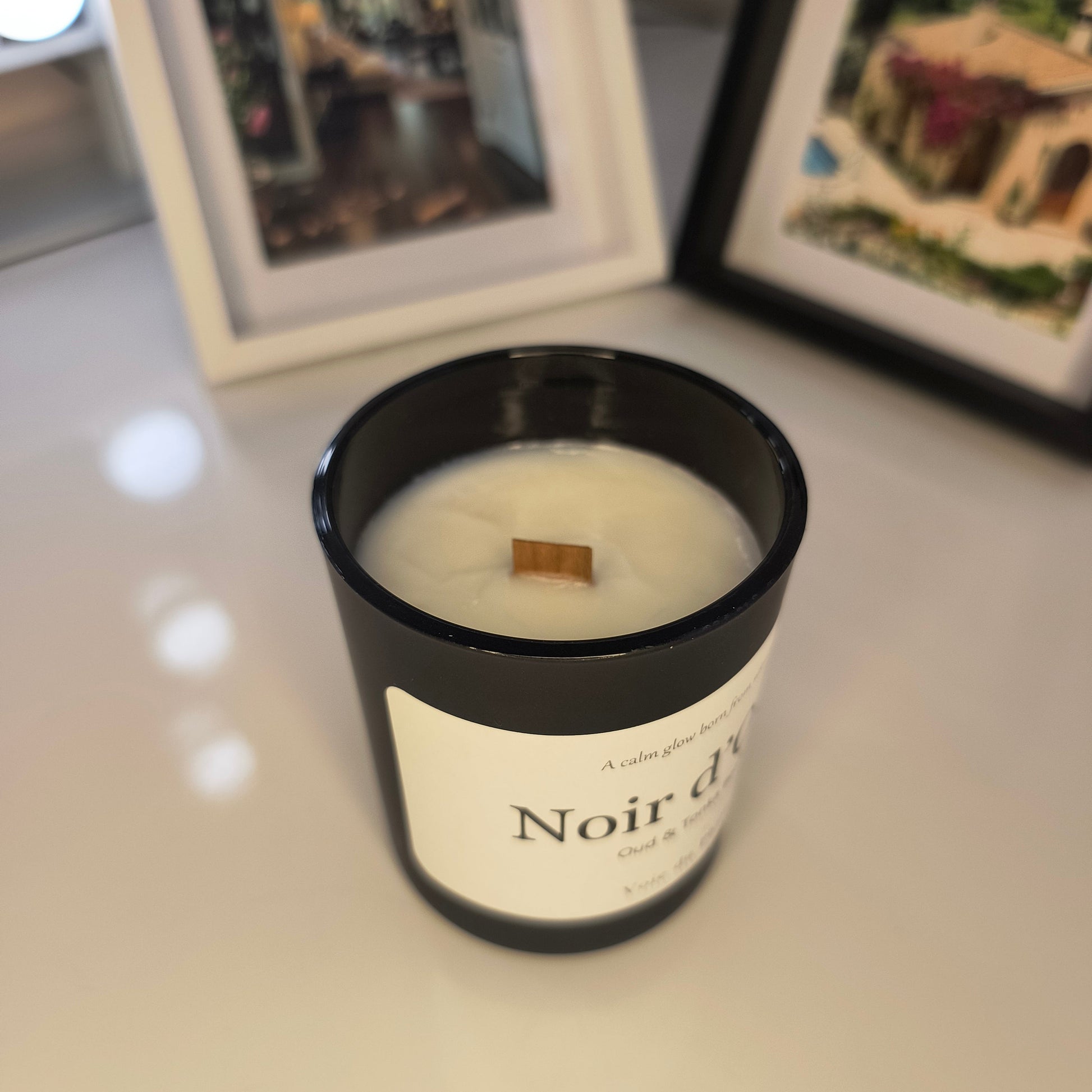 Candle in a black container with a white label on a surface, with framed pictures in the background.