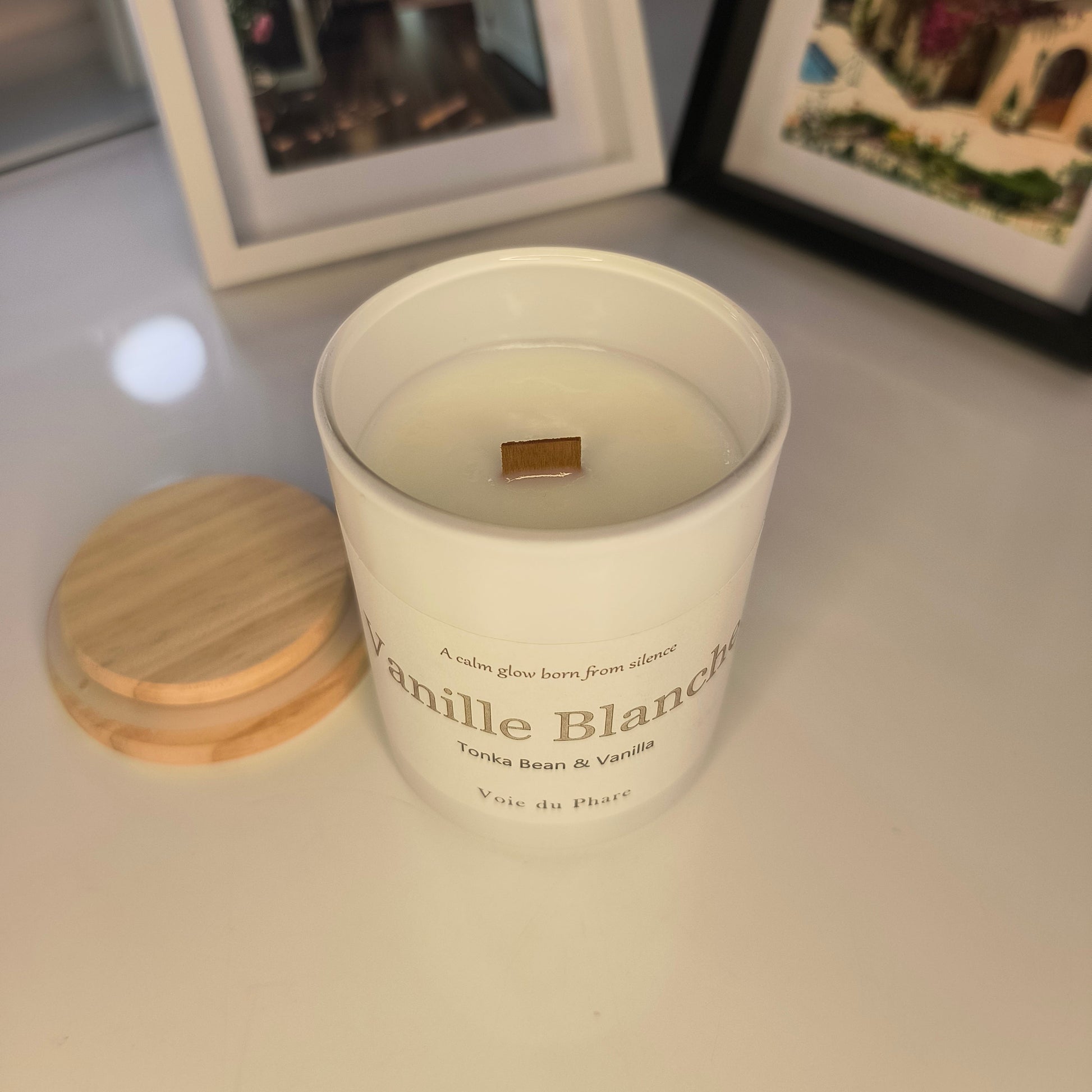 White candle in a wooden holder on a surface with framed pictures in the background